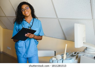Young african-american female doctor in white coat using ultra ultrasound scanning machine and looking on the screen. African woman doctor working on modern ultrasound equipment. - Shutterstock ID 1811281513
