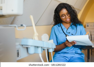 Young african-american female doctor in white coat using ultra ultrasound scanning machine and looking on the screen. African woman doctor working on modern ultrasound equipment. - Shutterstock ID 1811281522