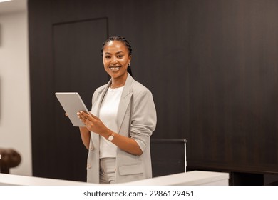 The young African-American female receptionist stands behind the reception desk with a digital tablet in her hands and smiles at the camera.