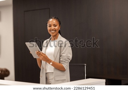 The young African-American female receptionist stands behind the reception desk with a digital tablet in her hands and smiles at the camera.
