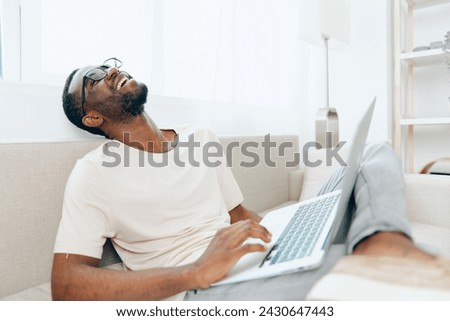 Young AfricanAmerican freelancer working on a laptop and typing happily while sitting on a modern sofa in a cozy living room With a casual and relaxed atmosphere, he embraces the benefits of education