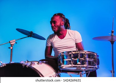 Young african-american jazz musician or drummer playing drums on blue studio background in trendy neon lights. Concept of music, hobby, inspirness. Colorful portrait of joyful attractive artist.