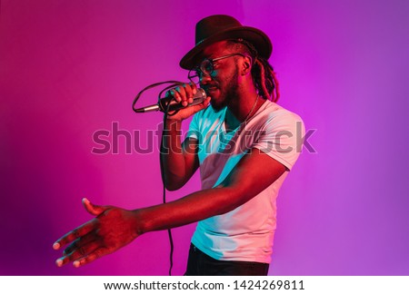 Young african-american jazz musician with microphone singing a song on purple studio background in trendy neon light. Concept of music, hobby, inspirness. Colorful portrait of joyful attractive artist