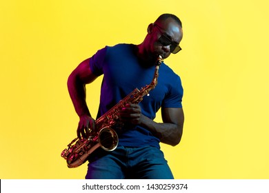 Young african-american jazz musician playing the saxophone on yellow studio background in trendy neon light. Concept of music, hobby. Joyful attractive guy improvising. Colorful portrait of artist.