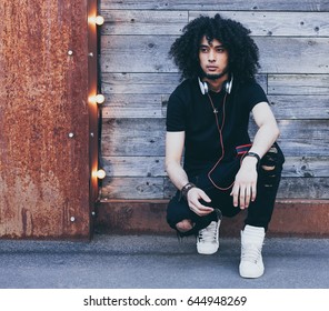 A Young African-American Man With An Afro Hairstyle Listens To Music As DJ, Resting, Sitting. Dressed In A Black Outfit. Streetstyle. Reggae. Outdoor.