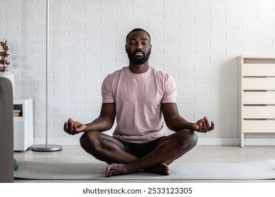 A young African-American man is meditating in a bright, minimalist living room, sitting cross-legged on the floor with eyes closed, practicing mindfulness and relaxation. - Powered by Shutterstock