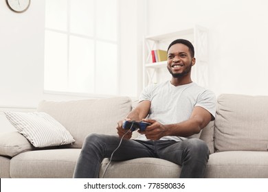 Young African-american Man Playing Video Games At Home. Happy Guy Sitting On The Couch With Joystick In Hands, Copy Space