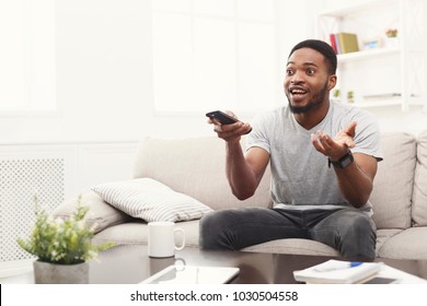 Young African-american Man Watching Tv. Emotional Guy Pointing With Remote Controller On Tv-set. Man Worrying About Favourite Football Team At Home, Copy Space