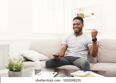 Young african-american man watching tv on the couch, happy of favourite football team, pointing with remote controller on tv-set, copy space - Powered by Shutterstock