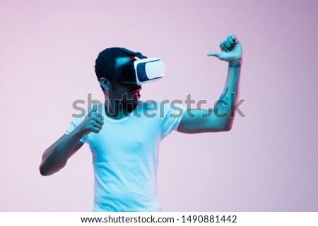 Young african-american man's pointing and using VR-glasses in neon light on gradient background. Male portrait. Concept of human emotions, facial expression, modern gadgets and technologies.