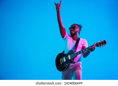 Young african-american musician playing the guitar like a rockstar on blue studio background in neon light. Concept of music, hobby. Joyful attractive guy improvising. Retro colorful portrait.