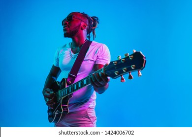 Young african-american musician playing the guitar like a rockstar on blue studio background in neon light. Concept of music, hobby. Joyful attractive guy improvising. Retro colorful portrait. - Shutterstock ID 1424006702