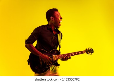 Young african-american musician playing the guitar like a rockstar on yellow background in neon light. Concept of music, hobby, festival, open-air. Joyful attractive guy improvising, singing song. - Powered by Shutterstock