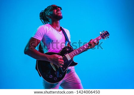 Young african-american musician playing the guitar like a rockstar on blue studio background in neon light. Concept of music, hobby. Joyful attractive guy improvising. Retro colorful portrait.
