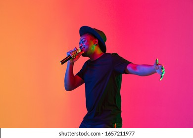 Young african-american musician singing on gradient orange-purple studio background in neon light. Concept of music, hobby, festival. Joyful party host, stand upper. Colorful portrait of artist.