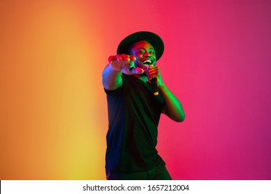 Young african-american musician singing on gradient orange-purple studio background in neon light. Concept of music, hobby, festival. Joyful party host, stand upper. Colorful portrait of artist. - Powered by Shutterstock