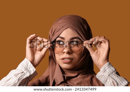 Young African-American Muslim woman in stylish sunglasses with beautiful makeup on brown background