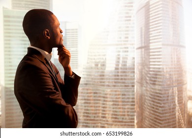 Young african-american thoughtful businessman wearing suit holding hand on chin, looking out of big office window at sunset city building lost in thoughts, planning future project. Side view copyspace