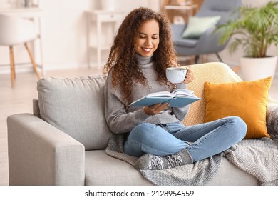 Young African-American woman with cup of tea reading book on sofa at home - Powered by Shutterstock