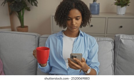 Young african-american woman with curly hair sitting in a living room, holding a red mug and using a smartphone. - Powered by Shutterstock