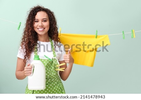 Young African-American woman with detergent, hanging laundry and clothespins showing thumb-up on color background