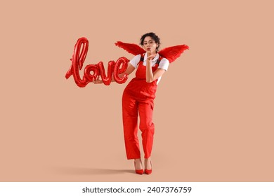 Young African-American woman dressed as Cupid with word LOVE made of balloons blowing kiss on beige background. Valentine's Day celebration