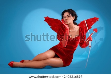 Young African-American woman dressed as Cupid with bow blowing kiss on blue background. Valentine's Day celebration