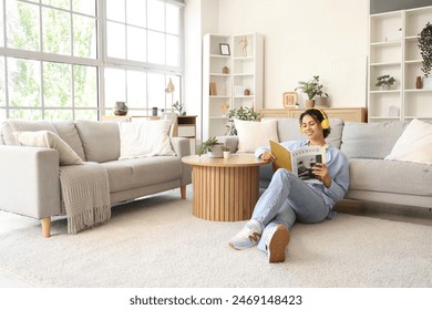 Young African-American woman in headphones reading magazine beside sofa at home - Powered by Shutterstock