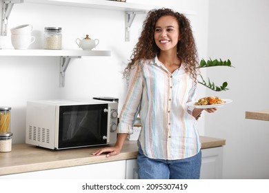 Joven afroamericana sosteniendo placa con comida cerca del horno microondas en la cocina