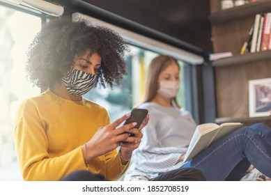 A Young African-American Woman Looks On Her Smart Phone At The Latest News Of Coronavirus Infection, Two Students In A Bookstore With Face Masks To Prevent The Contagion Of Covid19, Sun Flare Effect