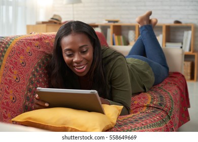 Young African-American Woman Lying On Sofa And Watching Comedy Movie On Tablet Computer