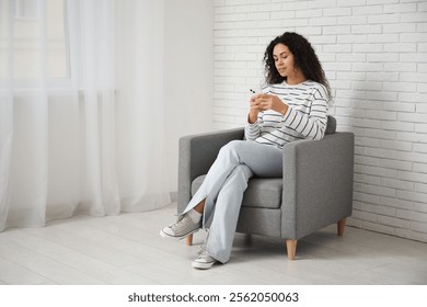 Young African-American woman with mobile phone sitting in grey armchair at home - Powered by Shutterstock