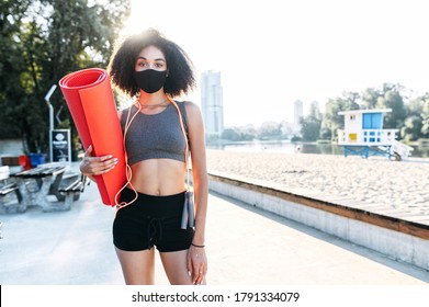 A Young African-American Woman With A Protective Medical Mask On The Face Going To Do Outdoor Workout. Biracial Girl With A Rolled Gymnastic Mat And Skipping Rope On The Waterfront