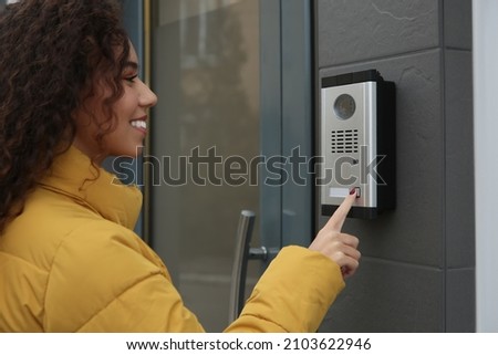 Young African-American woman ringing intercom with camera near building entrance