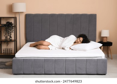 Young African-American woman in robe with pillow lying on soft mattress at home