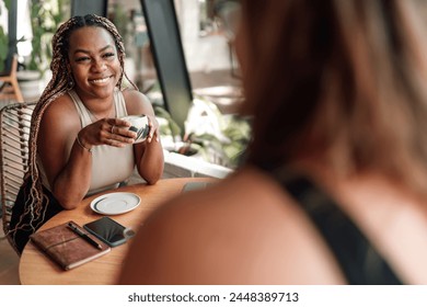 young African-American woman shares joyful moment with companion at cozy café. She's holding matcha cup, smiling warmly. table adorned with tech gadgets, suggesting modern, connected lifestyle - Powered by Shutterstock