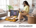 Young African-American woman unpacking suitcase with travelling accessories on sofa in hotel room