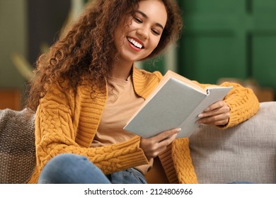 Young African-American woman in yellow cardigan reading book on sofa at home