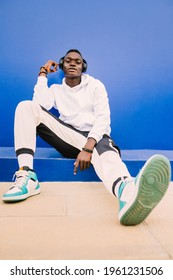 Young afro American black man wearing a white sweatshirt, basketball sneakers and headphones seated on a blue wall looking at camera with confidence