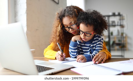 Young afro american child writing something in notebook and mother while working remotely from home laptop  mom helping small son and homework while sitting at desk at home  Childcare concept