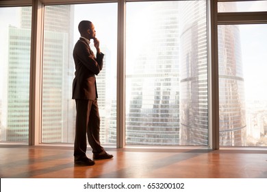 Young afro american contemplative businessman stands in office with hand on chin, looks through big window at the city, african entrepreneur enjoys sunset, rests after work, gets business inspiration
