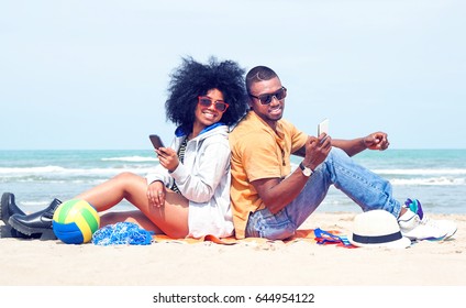 Young afro american couple having fun back to back at the beach using mobile phone - Attractive african models holding smartphone joyful summer moments sitting by ocean - Vintage filter modified ball