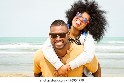 Young afro american couple playing piggyback ride on beach - Cheerful african friends having fun at day with blue ocean background - Concept of lovers happy moments on summer holiday - Vintage filter
