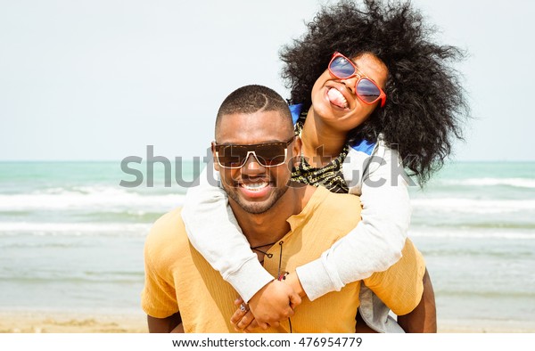 Young afro american couple playing piggyback ride\
on beach - Cheerful african friends having fun at day with blue\
ocean background - Concept of lovers happy moments on summer\
holiday - Vintage filter
