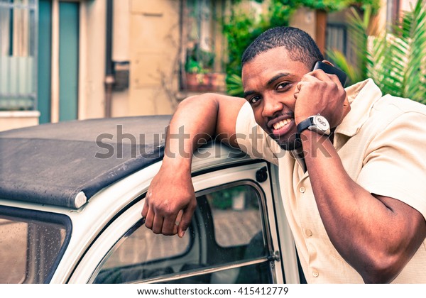 Young afro american guy speaking at mobile phone\
next old retro car - Black man smiling during phone conversation -\
Fashion with new trends concept - Soft focus on him - Vintage\
filtered look\
