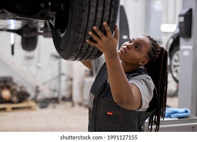 Young afro american mechanic adjusting the tire at repair garage alone, in overalls uniform, black woman working in auto repair shop, focused, raising tire up to lifted car. Side view