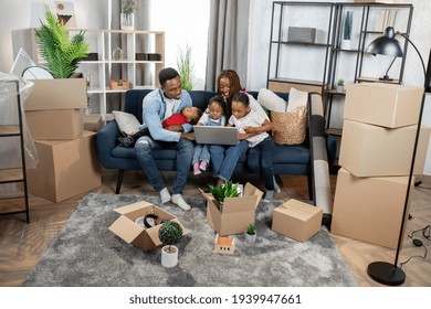 Young Afro American Parents With Their Three Kids Sitting Together On Couch And Using Modern Laptop. Happy Big Family Planning Design On New Apartment. Moving Boxes Around.