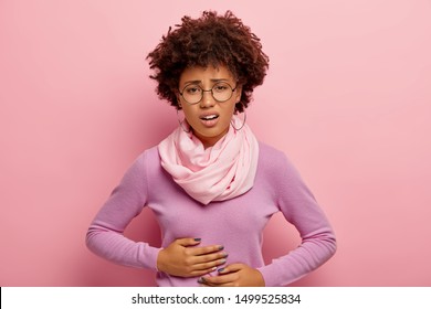 Young Afro American woman suffers from painful cramps in belly, has stomachache, frowns face from pain, wears round spectacles, purple jumper with scarf and earrings, being hungry, feels ache