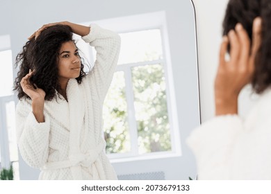 Young Afro American Woman Wearing In Bathrobe Look At Mirror, Making Daily Styling For Brunette Curly Hair. Female Standing In Bathroom At Home, Doing Haircare Treatment Procedure