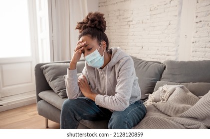 Young Afro American Woman Wearing Face Mask Feeling Ill, Exhausted And Sad On Sofa. Depressed African Americanyoung Adult Tired Of Stress, Tension And Isolation At Home. COVID And Mental Health.